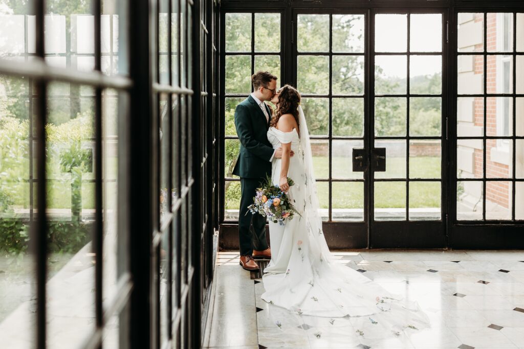 Bride and groom share a kiss in front of large black-framed windows at Gardencourt, with her floral-embroidered veil flowing across the marble floor.