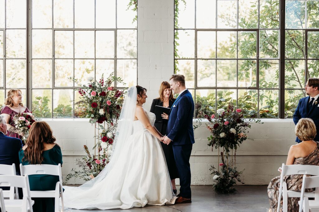 The Clerestory wedding ceremony with bride and groom exchanging vows in front of tall industrial windows and floral arrangements.
