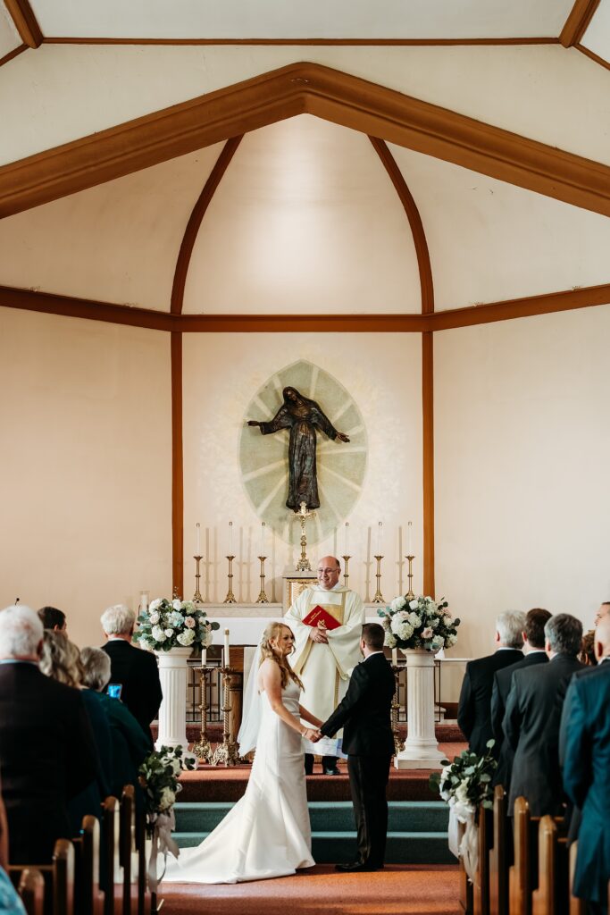 Hubers orchard winery wedding ceremony at St Marys of the Knobs as the bride and groom hold hands at the altar surrounded by guests.