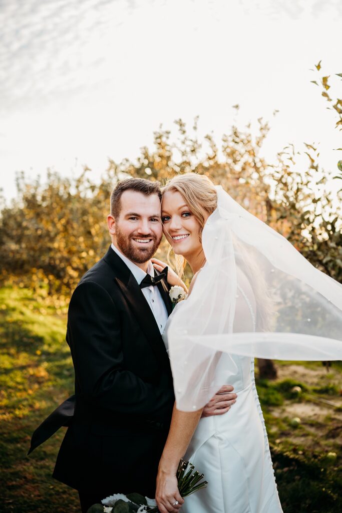Bride and groom portrait in orchard at Hubers Orchard and Winery with veil flowing in soft sunset light.