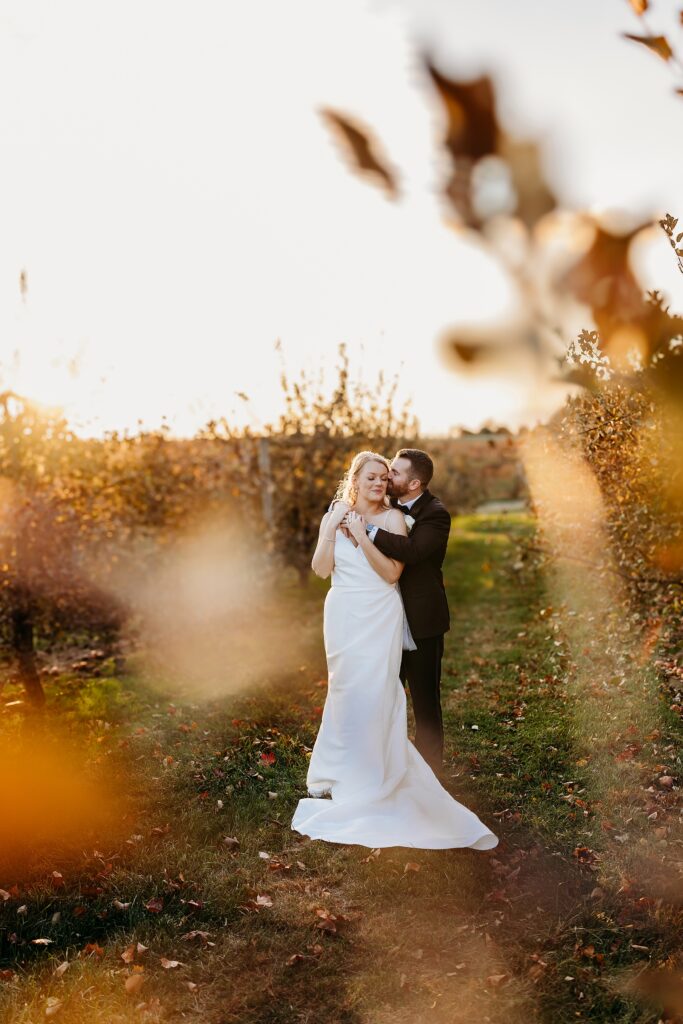 Bride and groom embrace in orchard at Hubers Orchard and Winery during golden hour with soft leaves framing the scene.