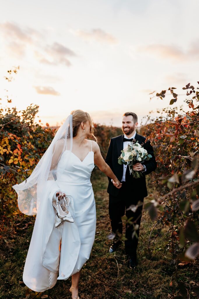 Bride and groom walk through orchard at Hubers Orchard and Winery at sunset with bride holding her shoes and groom carrying bouquet.