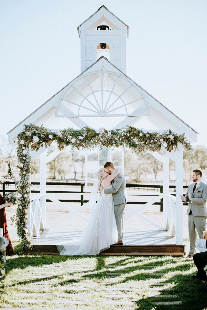 Barn at Merefield wedding ceremony with bride and groom kissing beneath white chapel altar and floral greenery arch.
