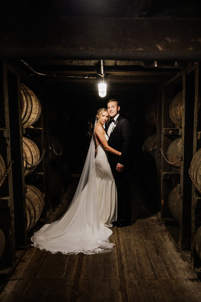 Castle & Key wedding couple posing in a historic bourbon barrel rickhouse surrounded by aging whiskey barrels.