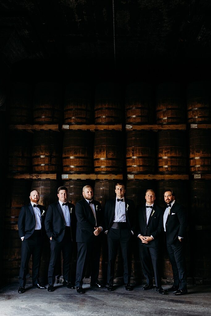 Castle & Key groomsmen portrait standing in front of stacked whiskey barrels inside the distillery rickhouse.