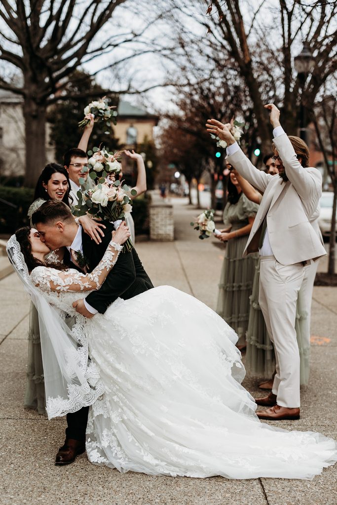 Bride and groom kiss while their wedding party cheers around them.