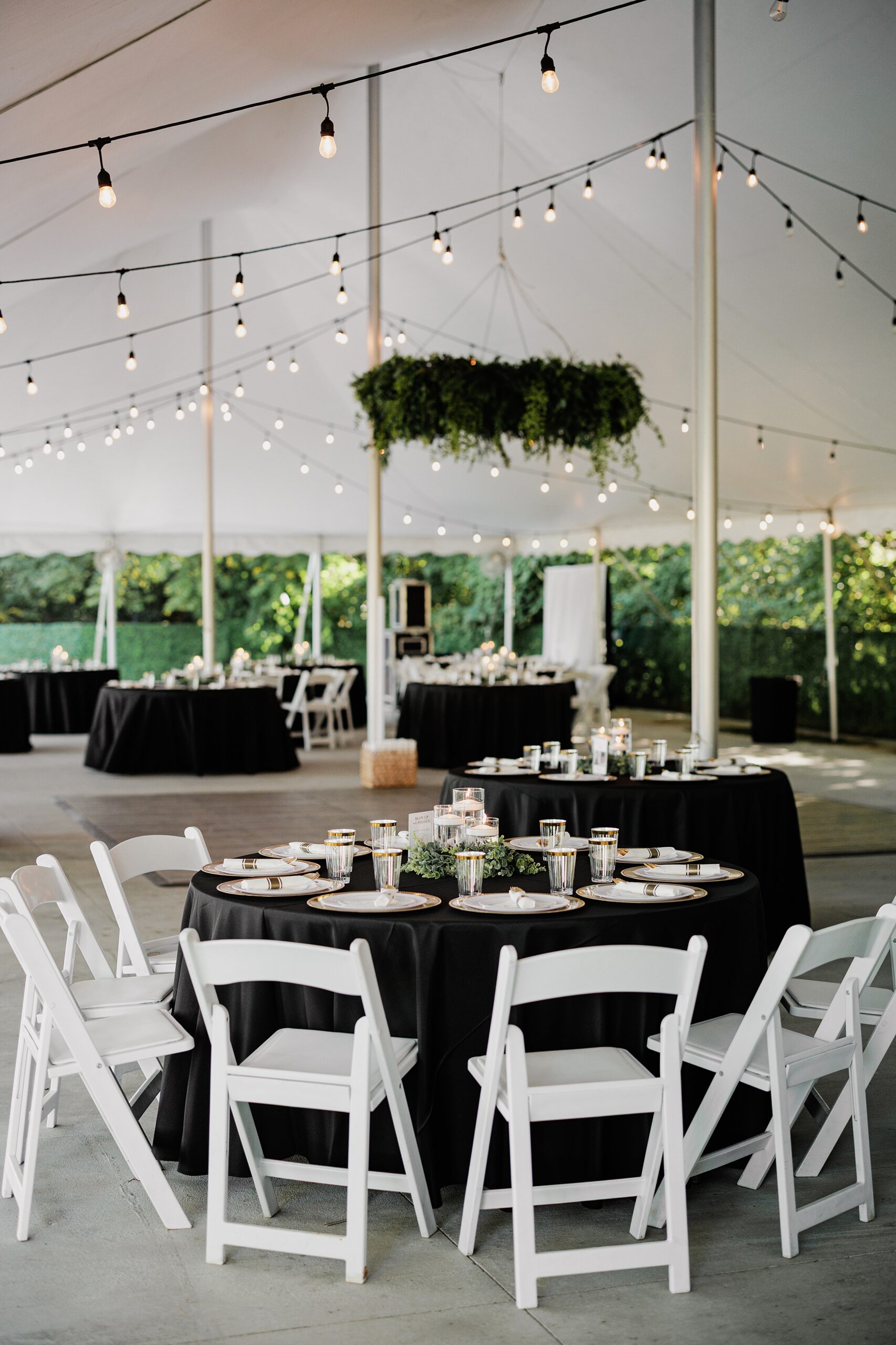 Tented wedding reception with black linens, white chairs, greenery and string lights.