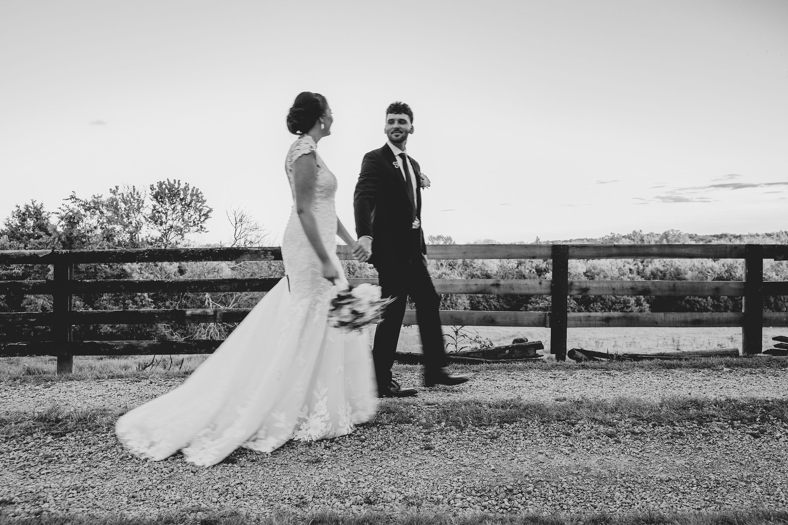 Black and white photo of a bride and groom walking hand in hand beside a fence.