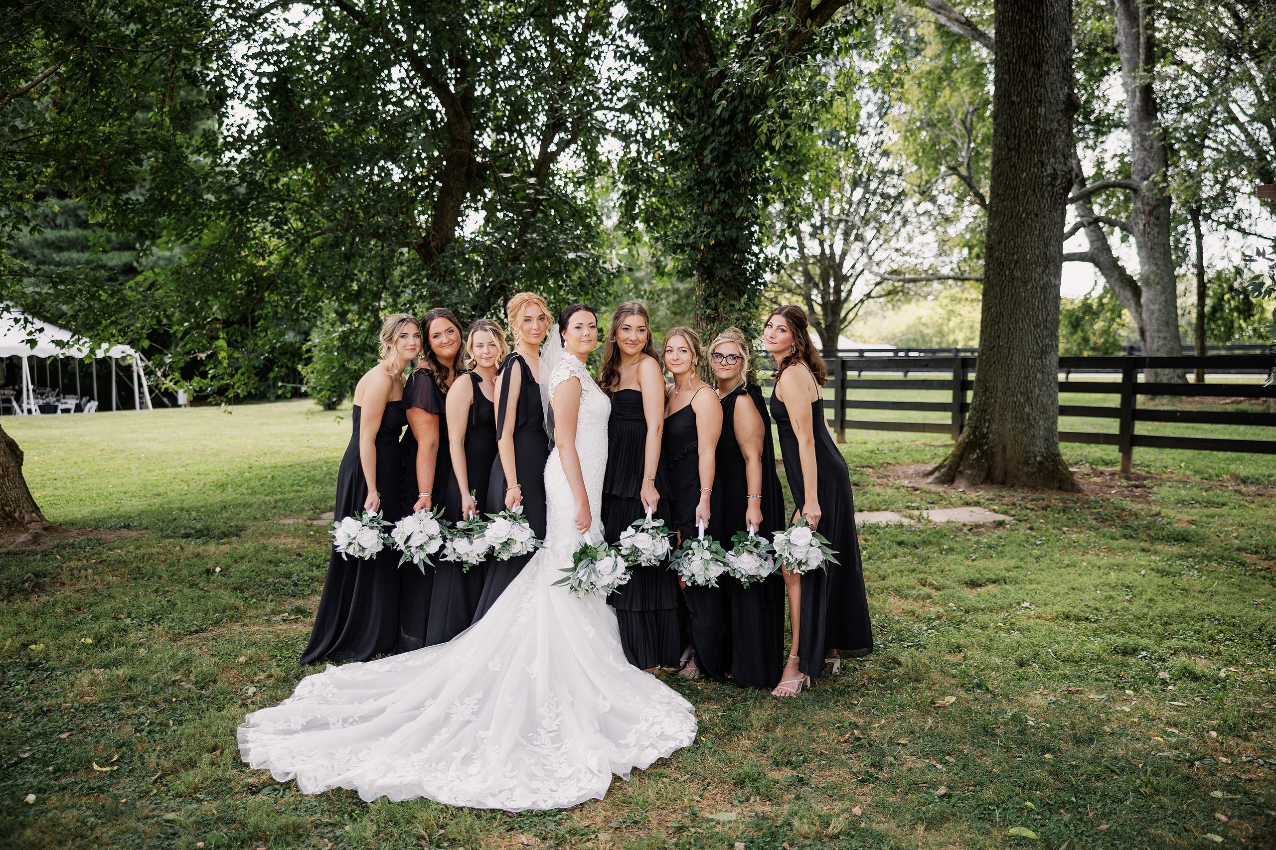 Bride and bridesmaids pose together beneath mature trees with coordinated white and green bouquets.