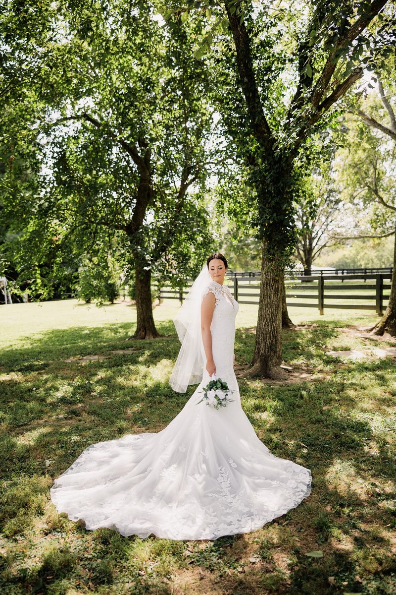 Bride posing beneath leafy trees with her bouquet and lace wedding dress train.