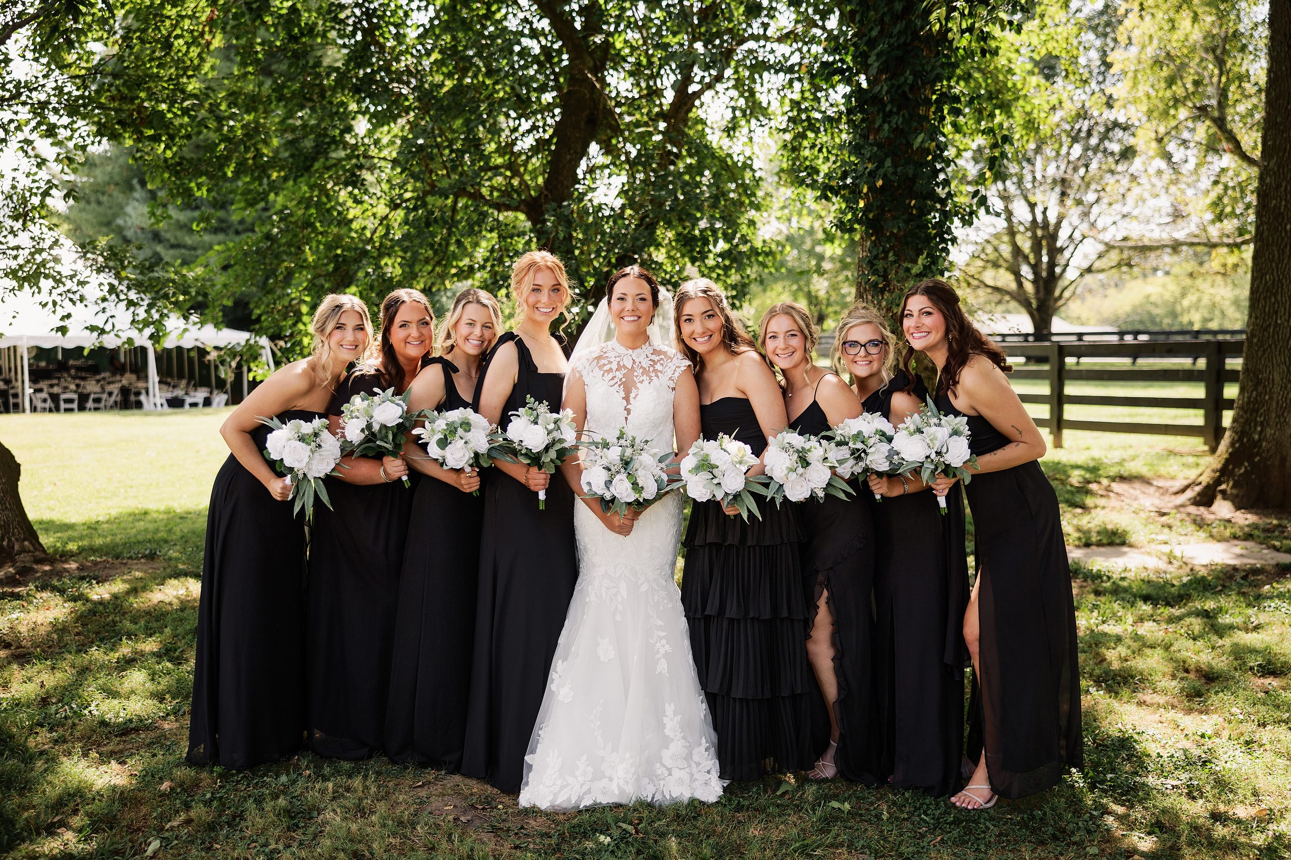 Bride poses with her bridesmaids in black dresses and white floral bouquets beneath the trees.
