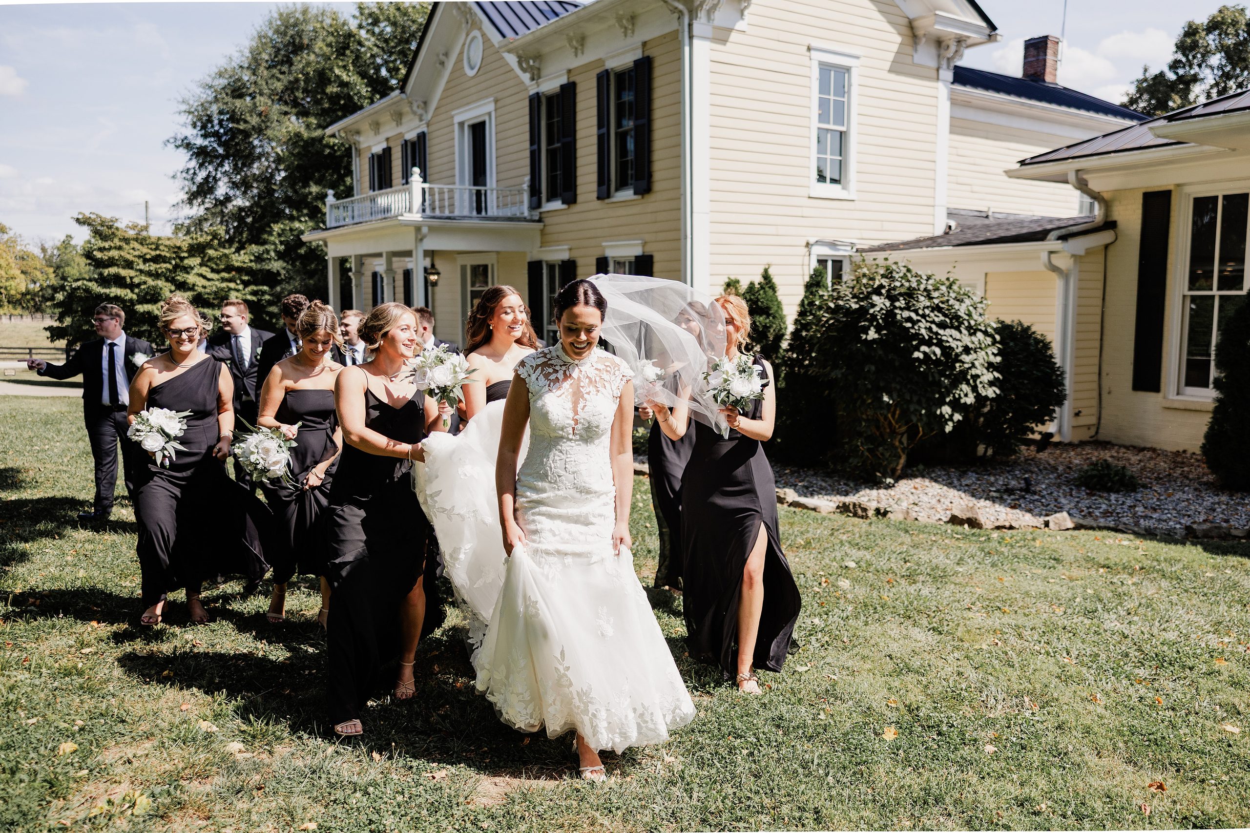 Bride and bridesmaids walk together outside in black dresses carrying white bouquets.