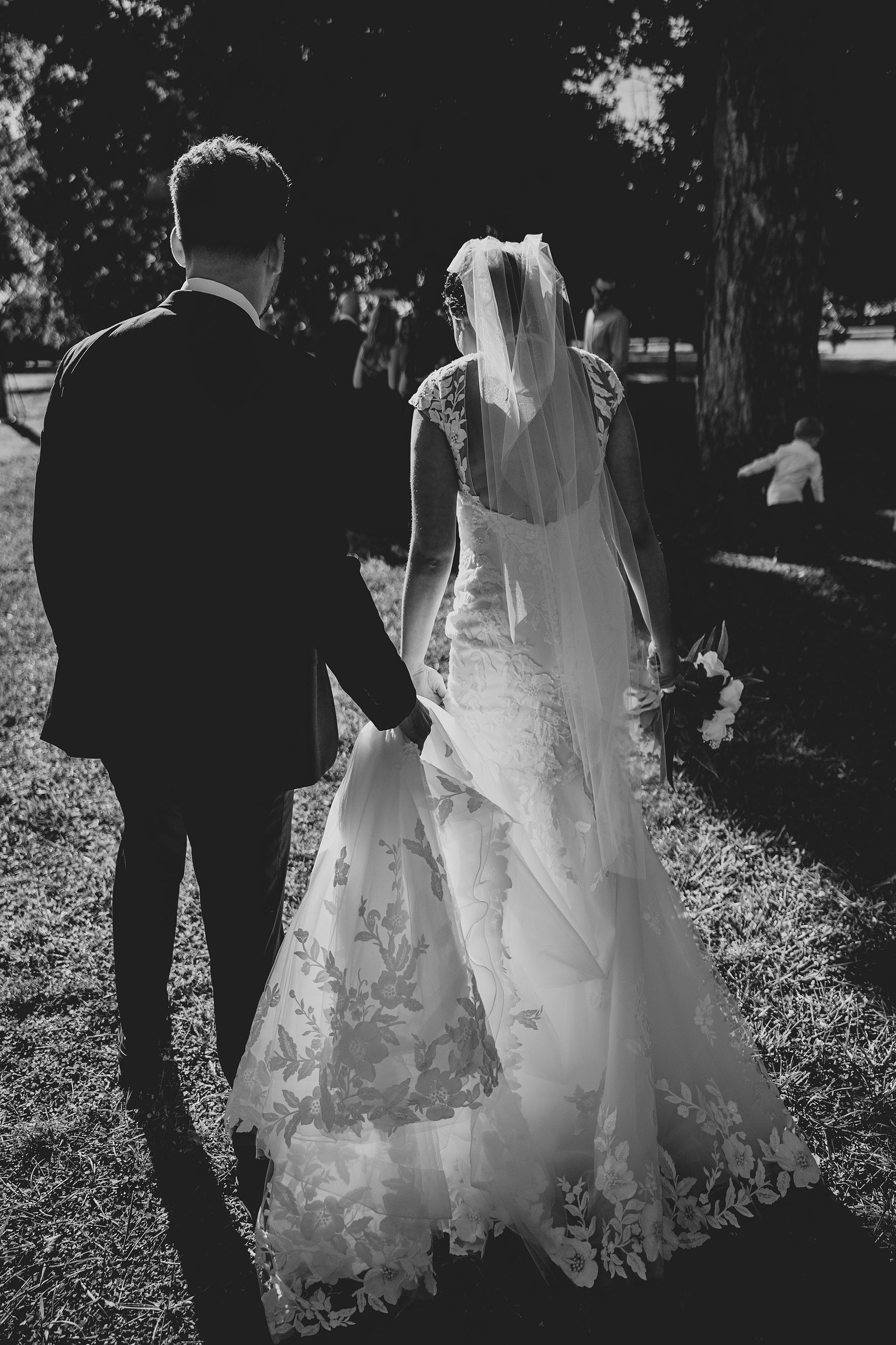Black and white candid of the bride and groom walking together beneath the trees.