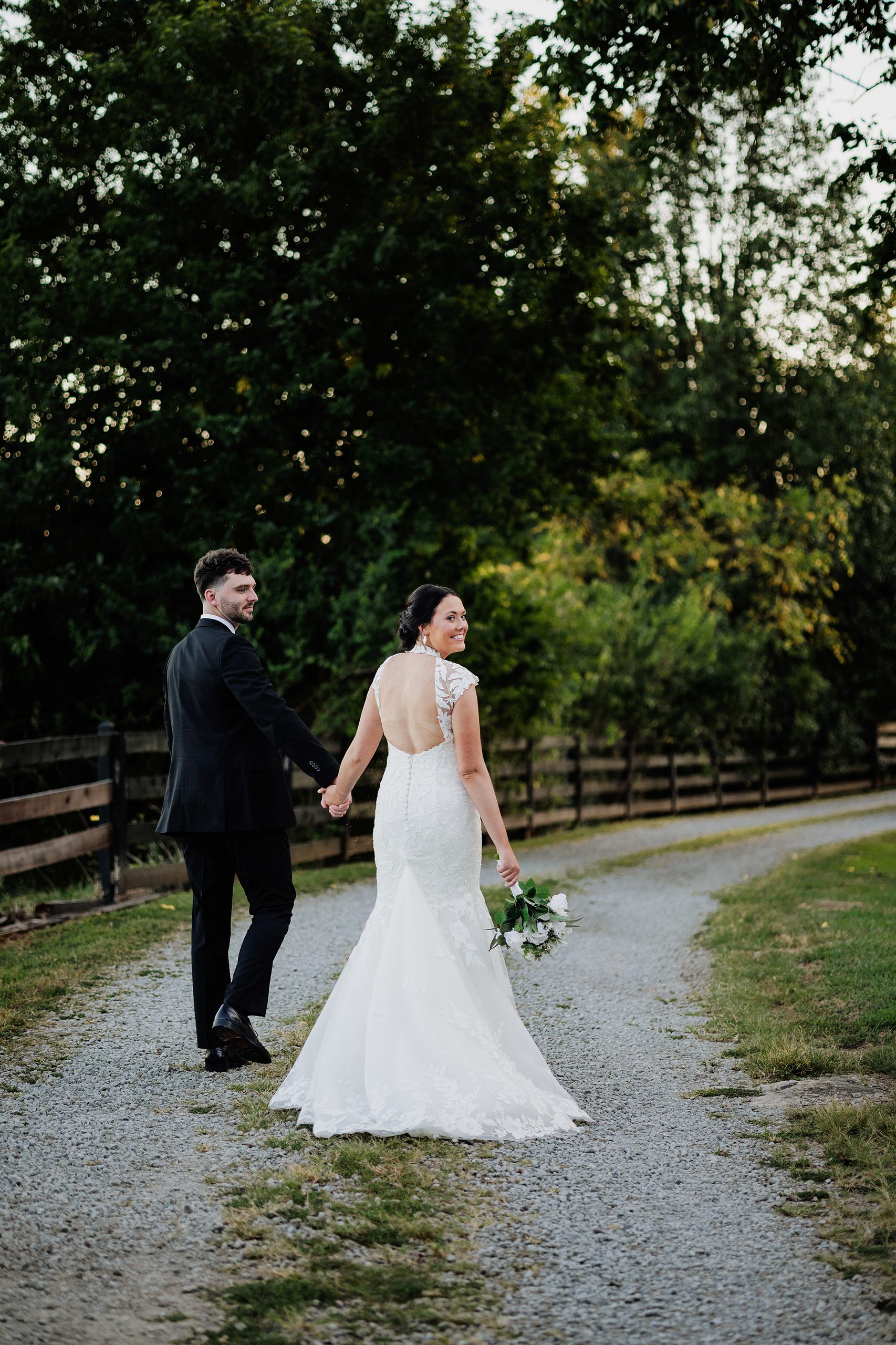 Bride and groom walk hand in hand along a tree-lined gravel lane.