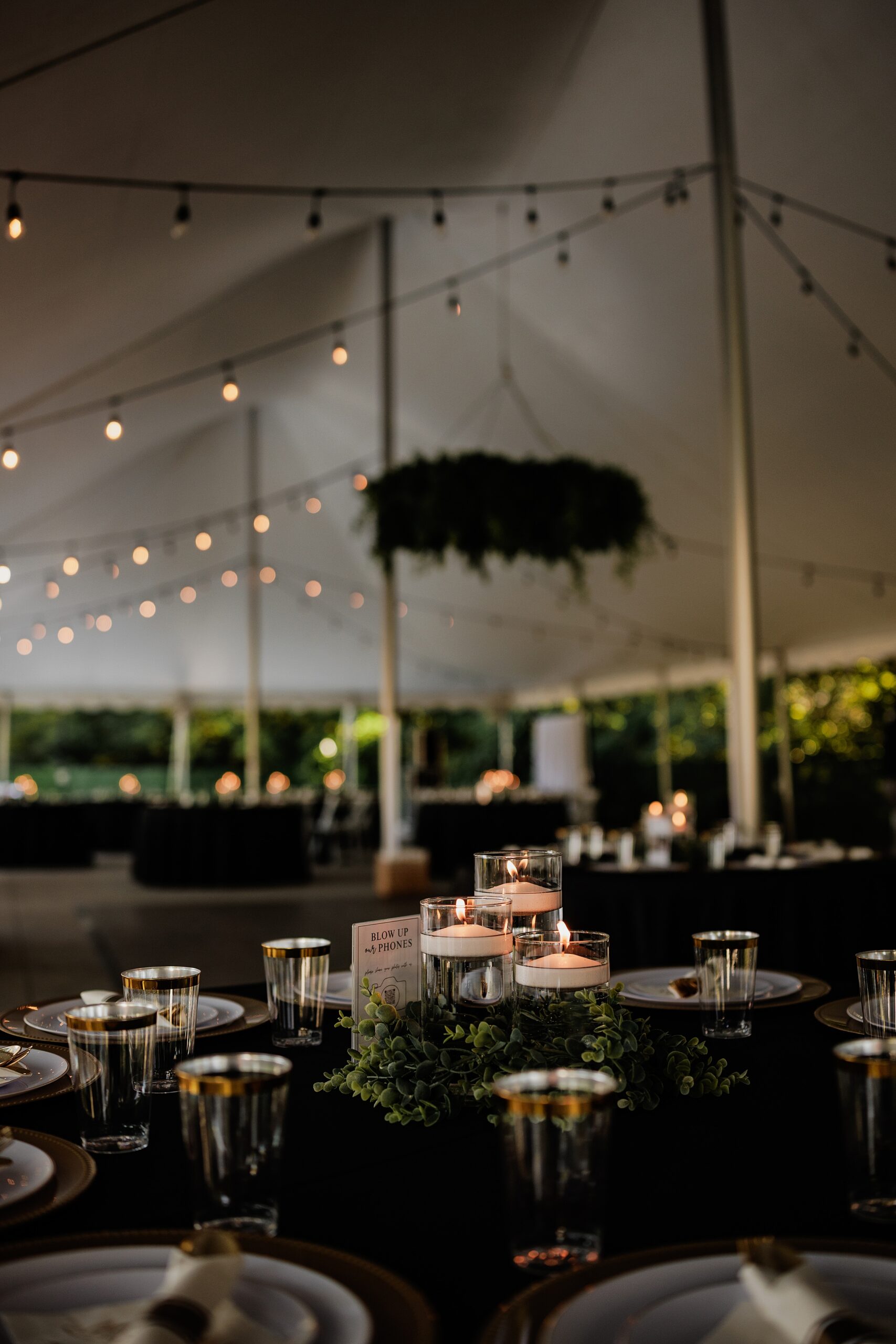Candlelit wedding reception table with greenery centerpiece, black linens, and gold-rimmed glassware.