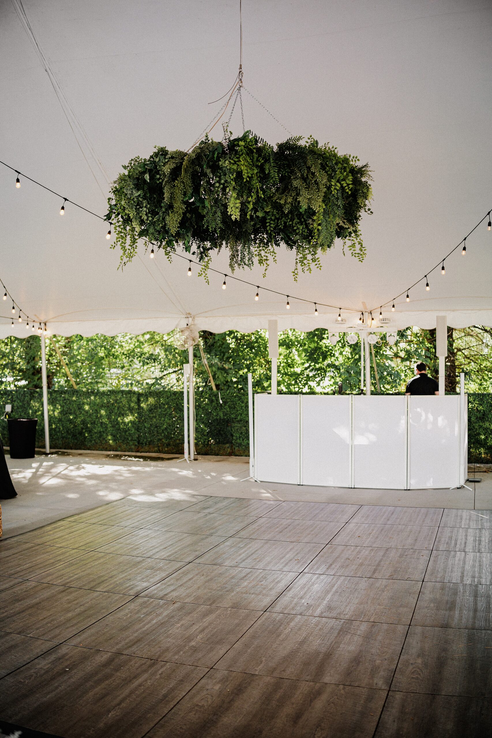 Wedding reception dance floor beneath hanging greenery and string lights inside a white tent.