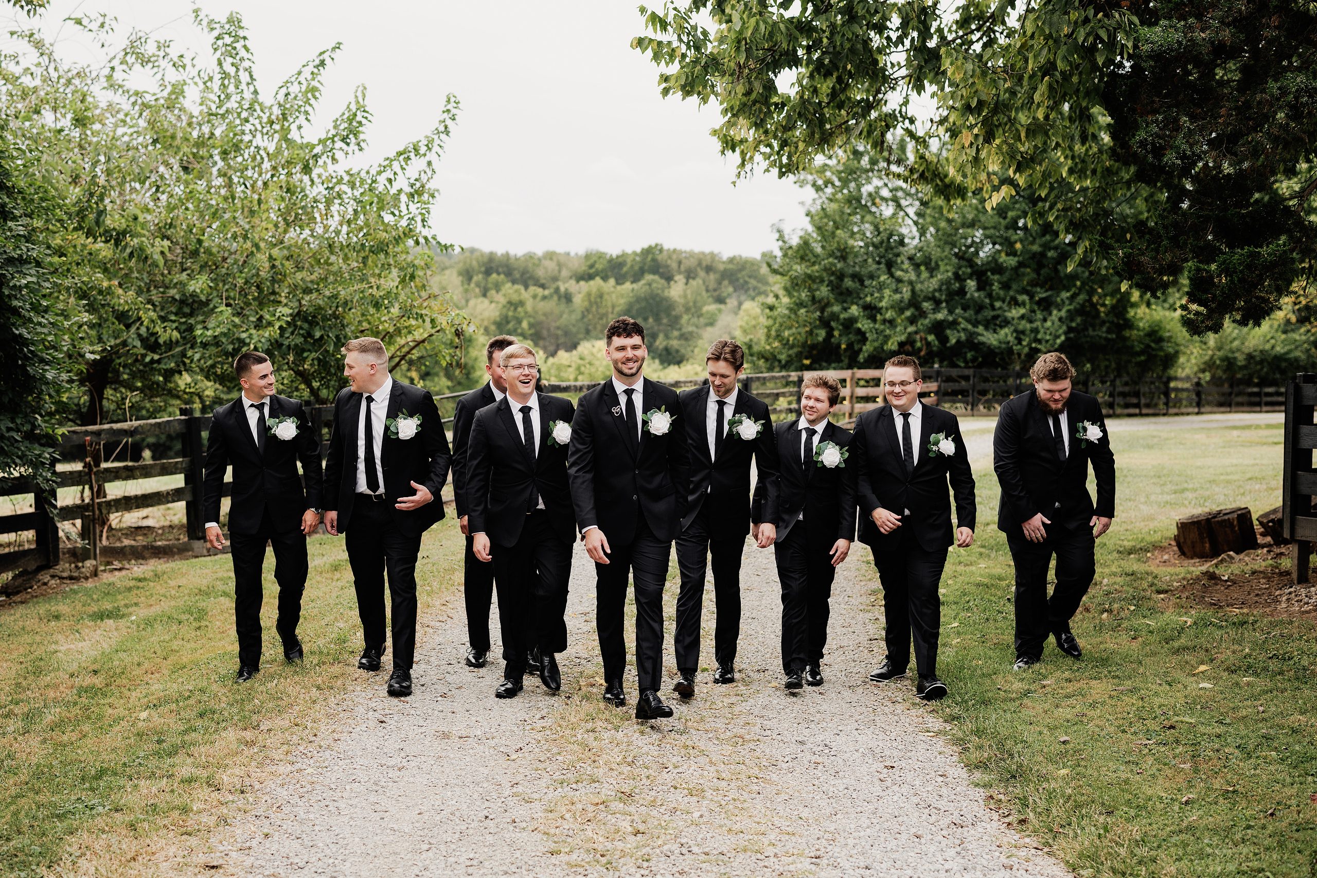 Groom and groomsmen walk together along a gravel path in matching black suits.
