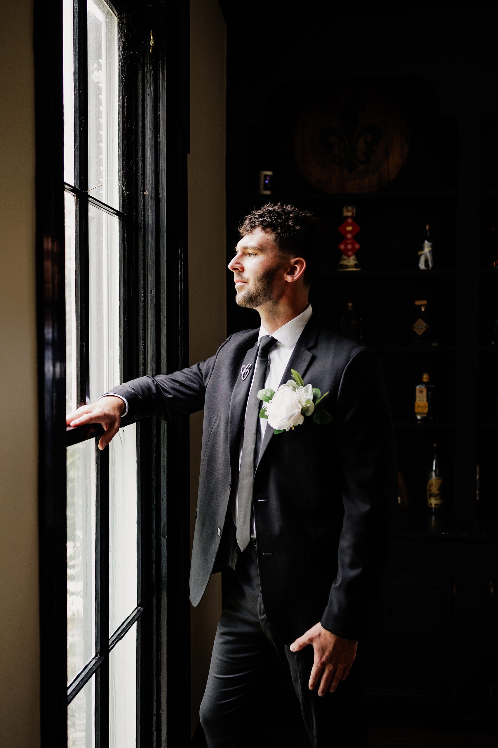 Groom in a black suit stands beside a window in dramatic natural light.