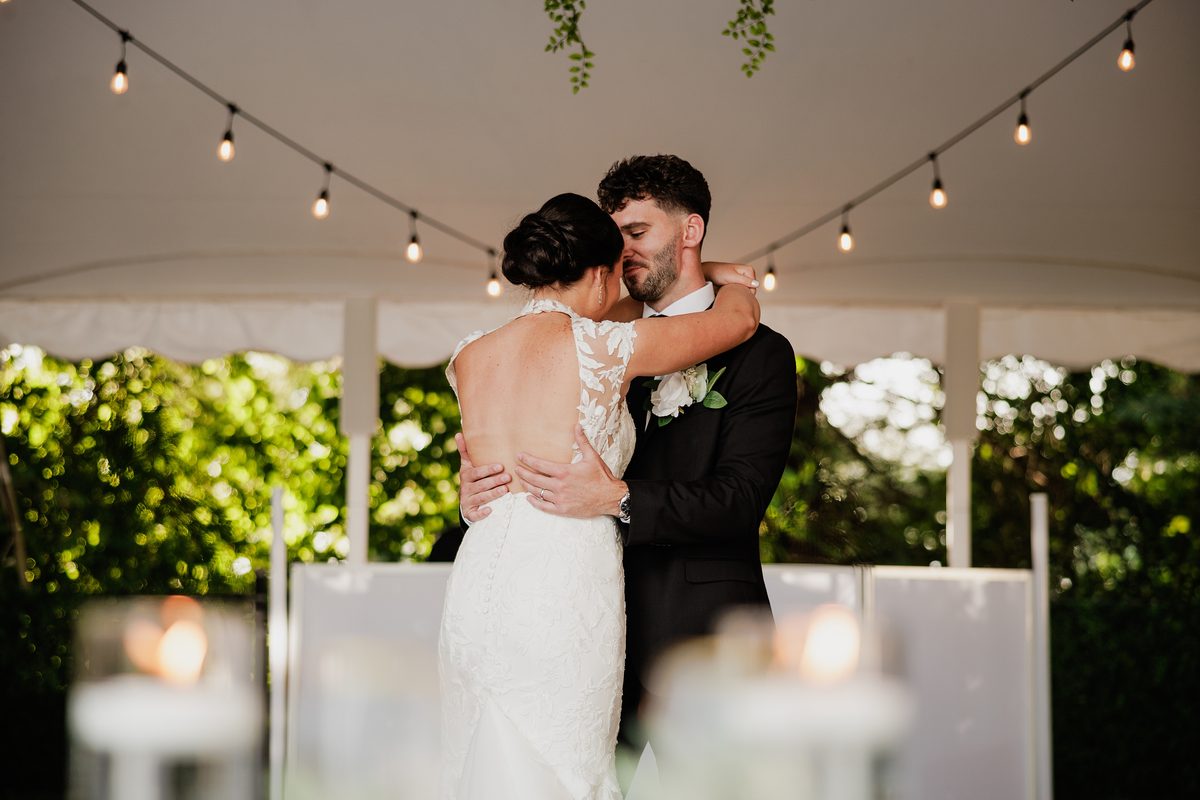 Newlyweds embrace during their first dance beneath string lights at their wedding reception.