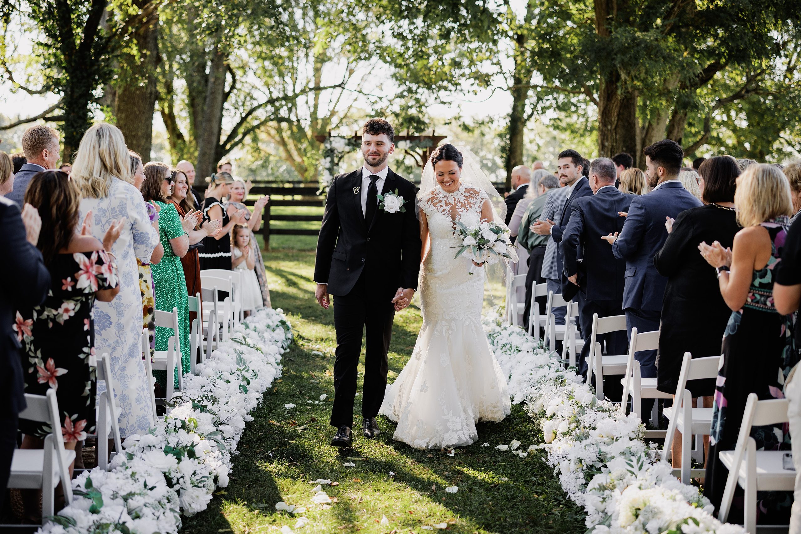 Outdoor wedding ceremony setup beneath mature trees with white chairs and floral-lined aisle.