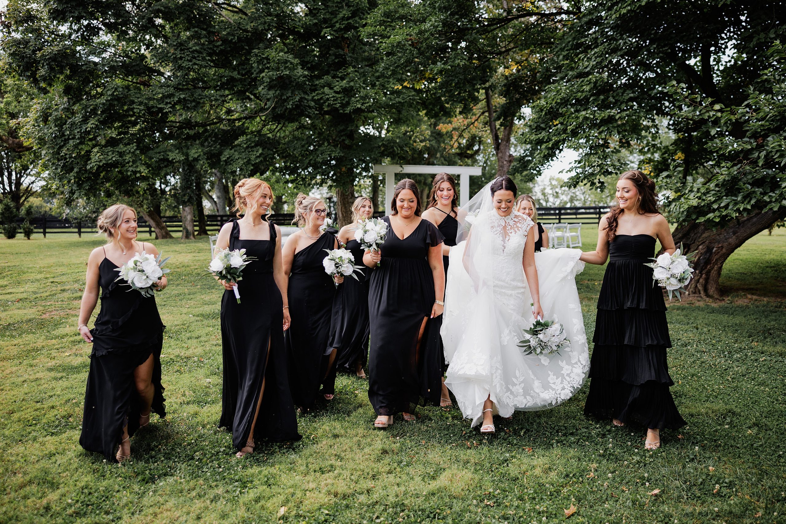 Bride and bridesmaids walk together outdoors carrying white bouquets in coordinating black dresses.