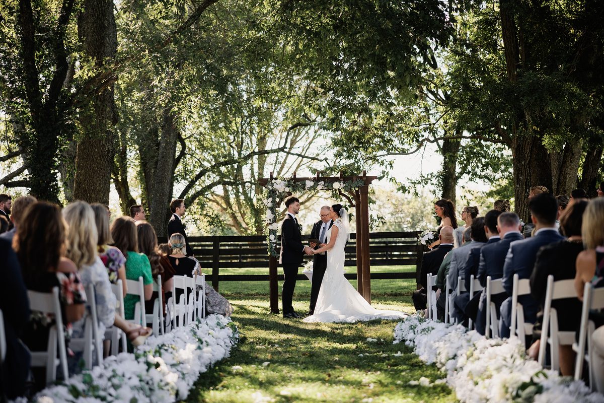 Couple exchanges vows beneath a floral arbor during their shaded outdoor wedding ceremony.