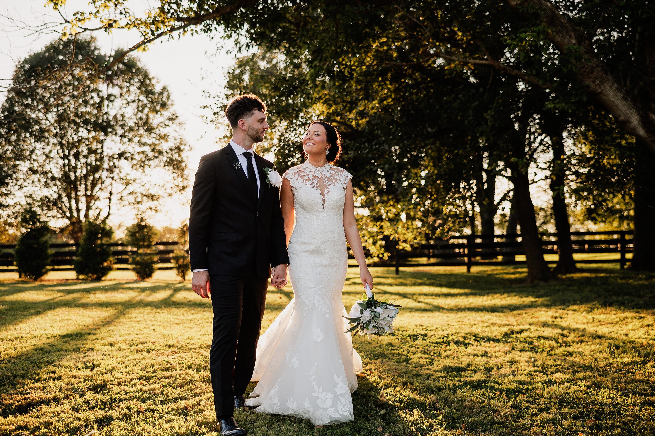 Bride and groom walk hand in hand through warm sunset light beneath mature trees.