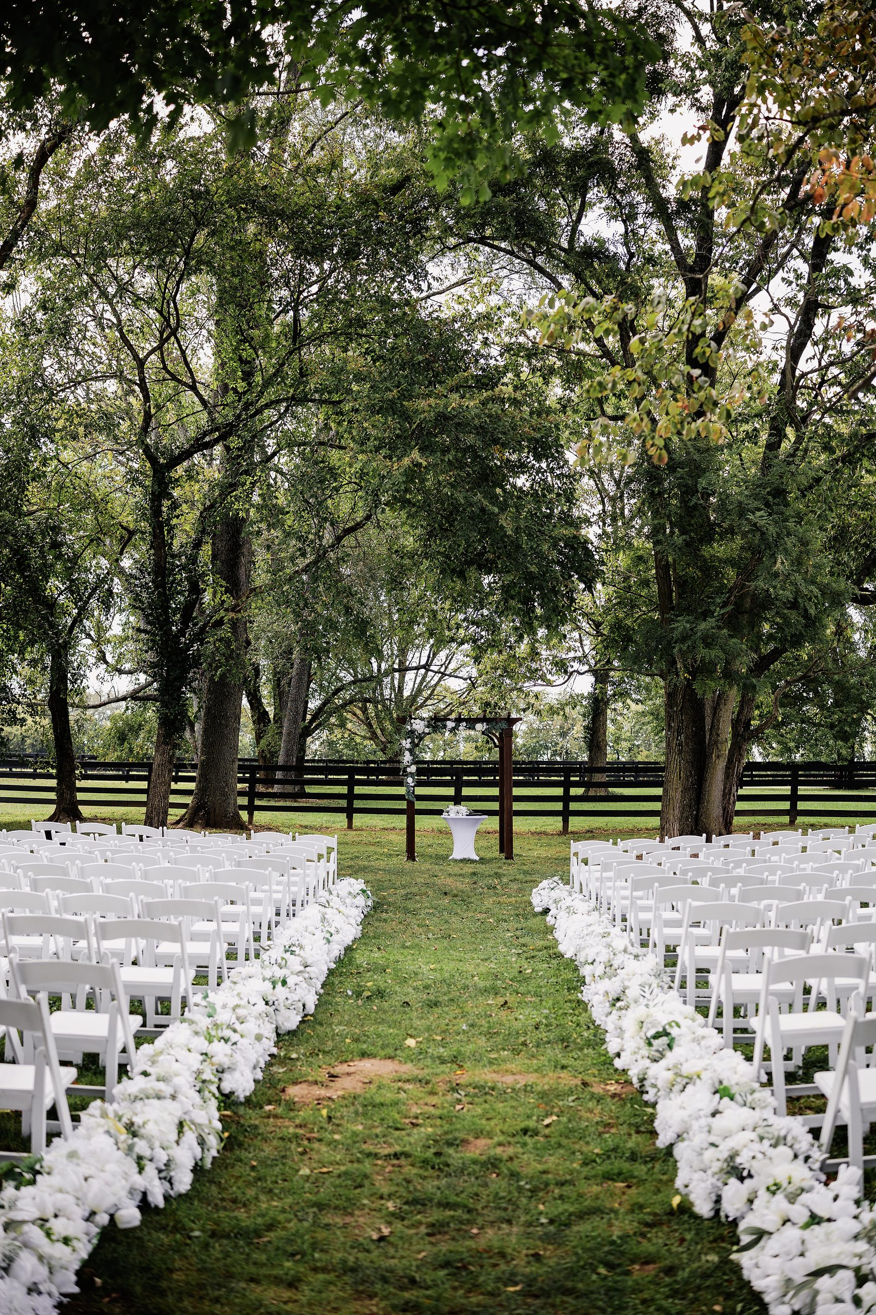 Outdoor wedding ceremony setup beneath mature trees with white chairs and floral-lined aisle.