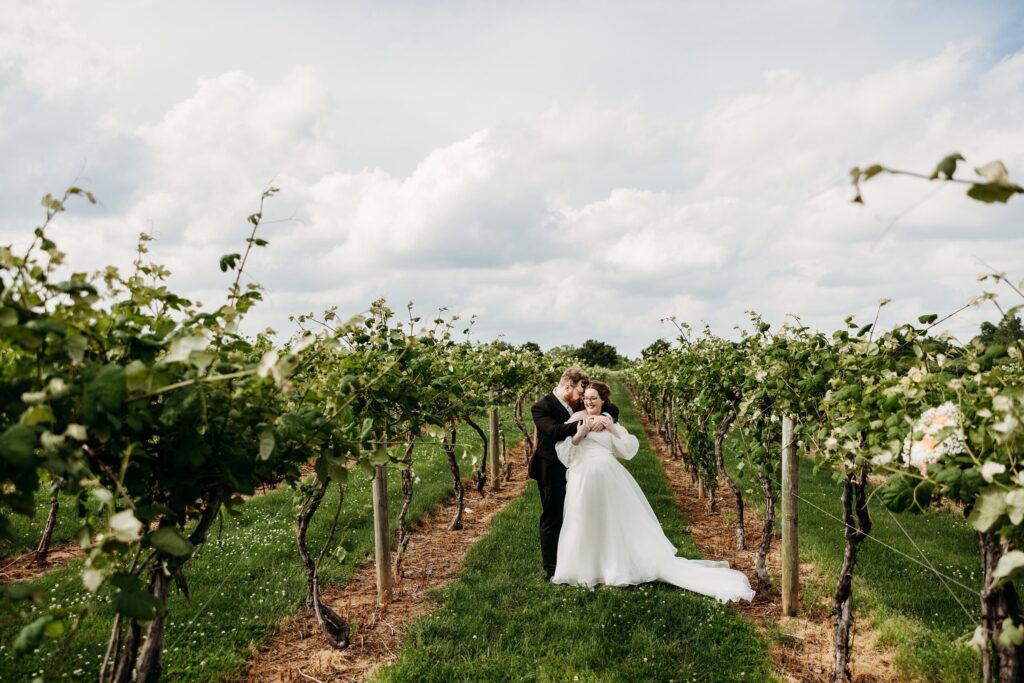 Groom embraces bride from behind in vineyard at Huber's Winery spring wedding.