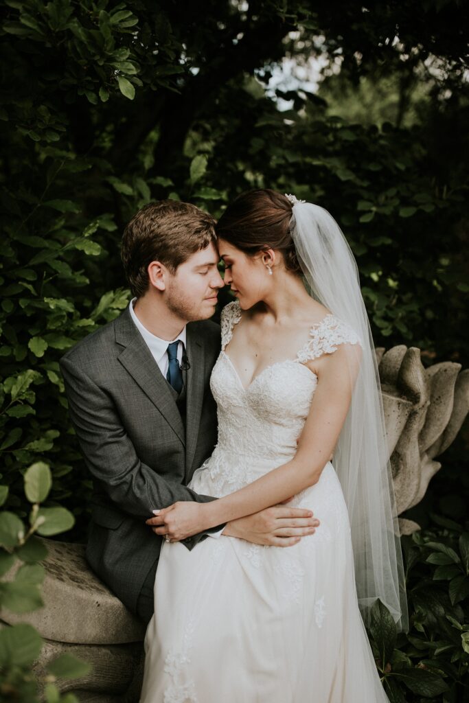 Yew Dell wedding couple portrait with the bride and groom embracing on a stone bench surrounded by lush garden greenery.