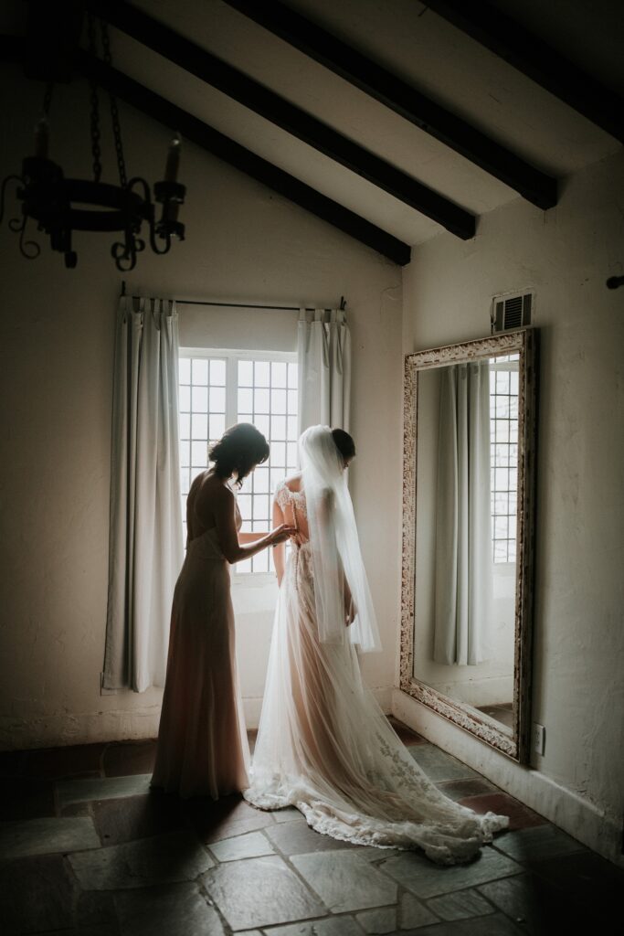 Yew Dell bride getting ready as her sister buttons the back of her wedding dress in front of a window.