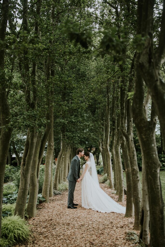 Yew Dell wedding portrait of the bride and groom standing beneath a tree lined garden path surrounded by lush greenery.