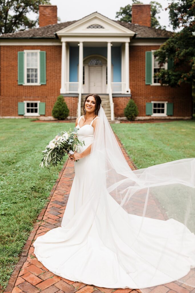 A bride poses with her bouquet in front of a historic home in Louisville, with her veil flowing behind her.
