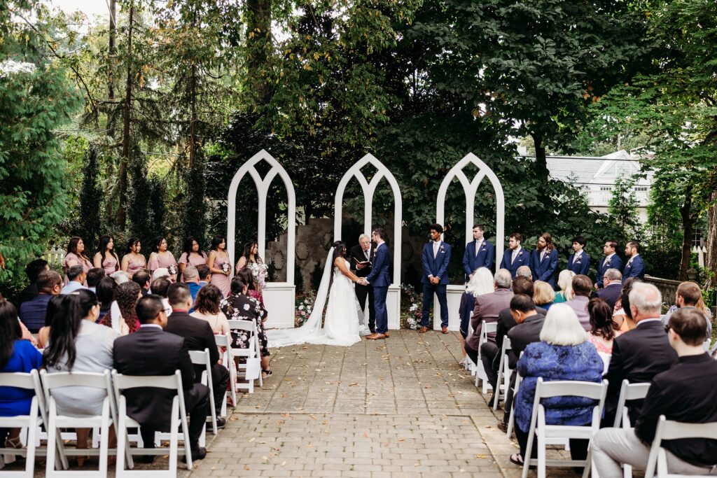 Intimate outdoor wedding ceremony in the courtyard with white arches, surrounded by mature trees and seated guests.