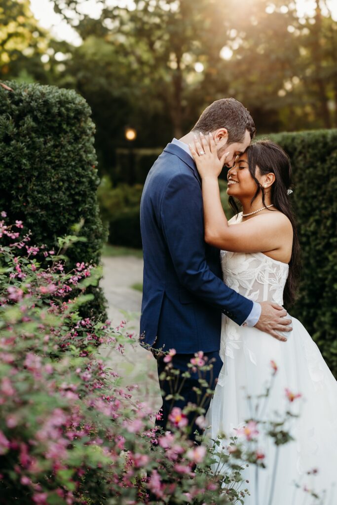 Bride and groom embracing among blooming flowers and manicured gardens during golden hour.