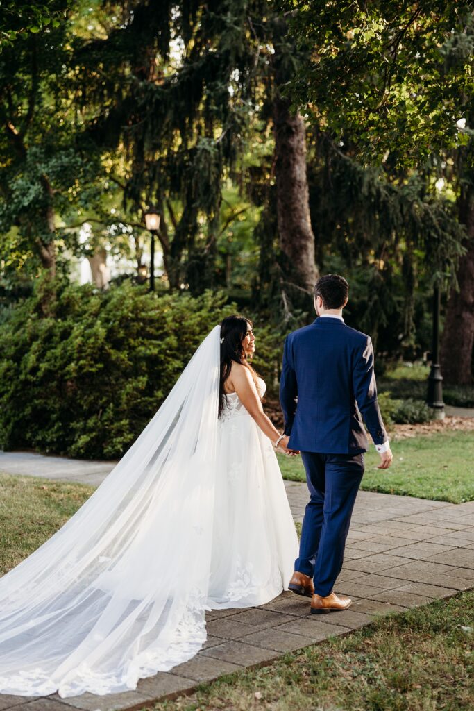 Bride and groom walking hand in hand along a garden path during golden hour with the bride’s veil flowing behind her.