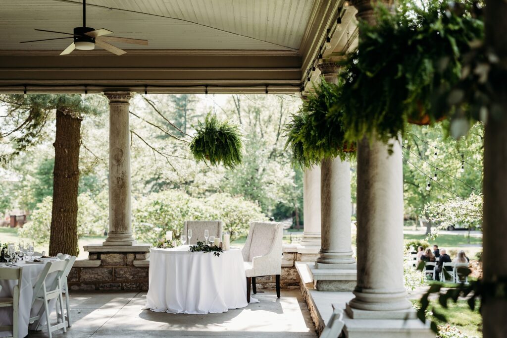 Covered patio set for an intimate wedding reception with sweetheart table, stone columns, and hanging ferns.
