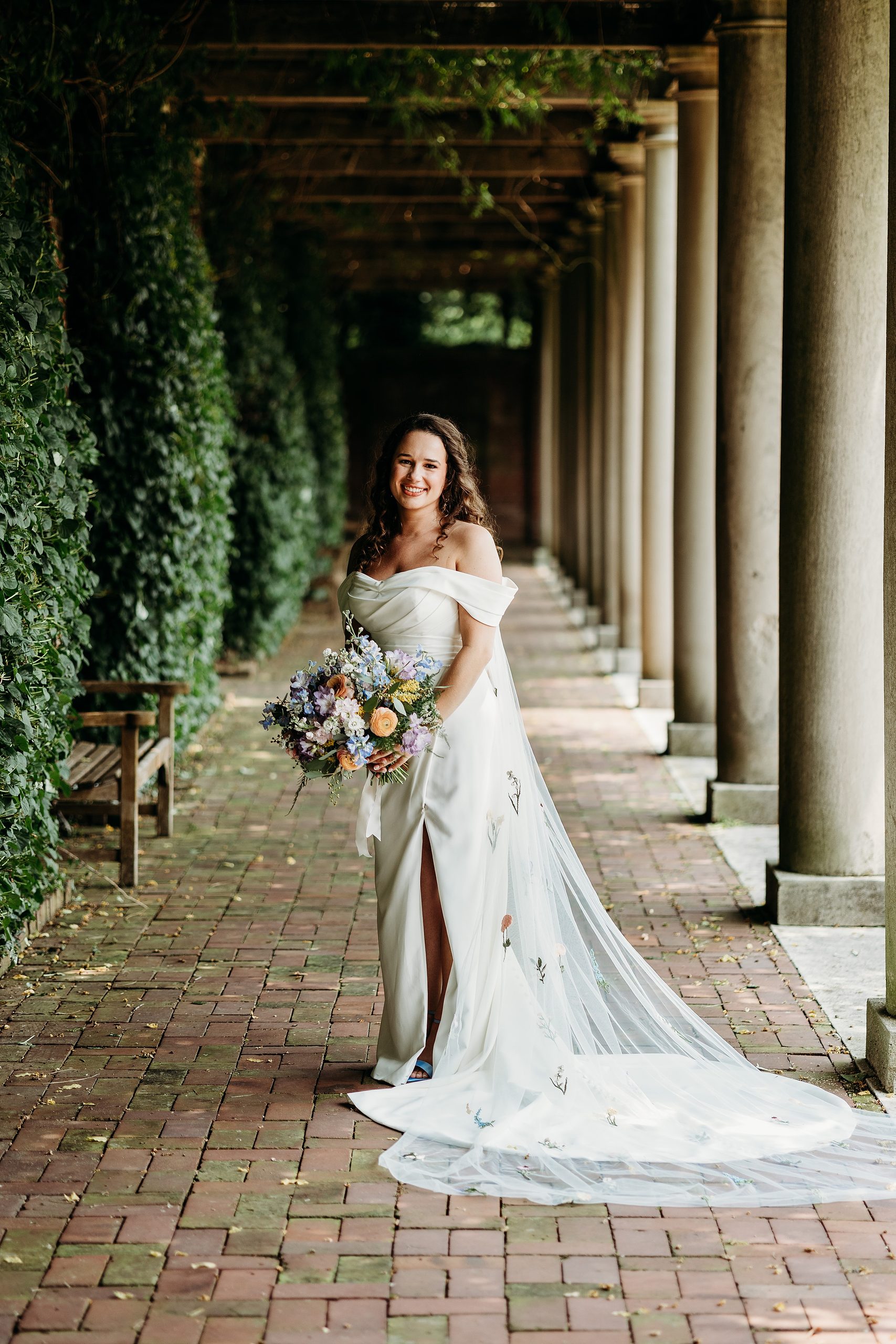 Bride with veil and bouquet stands on brick pathway next to greenery and columns.