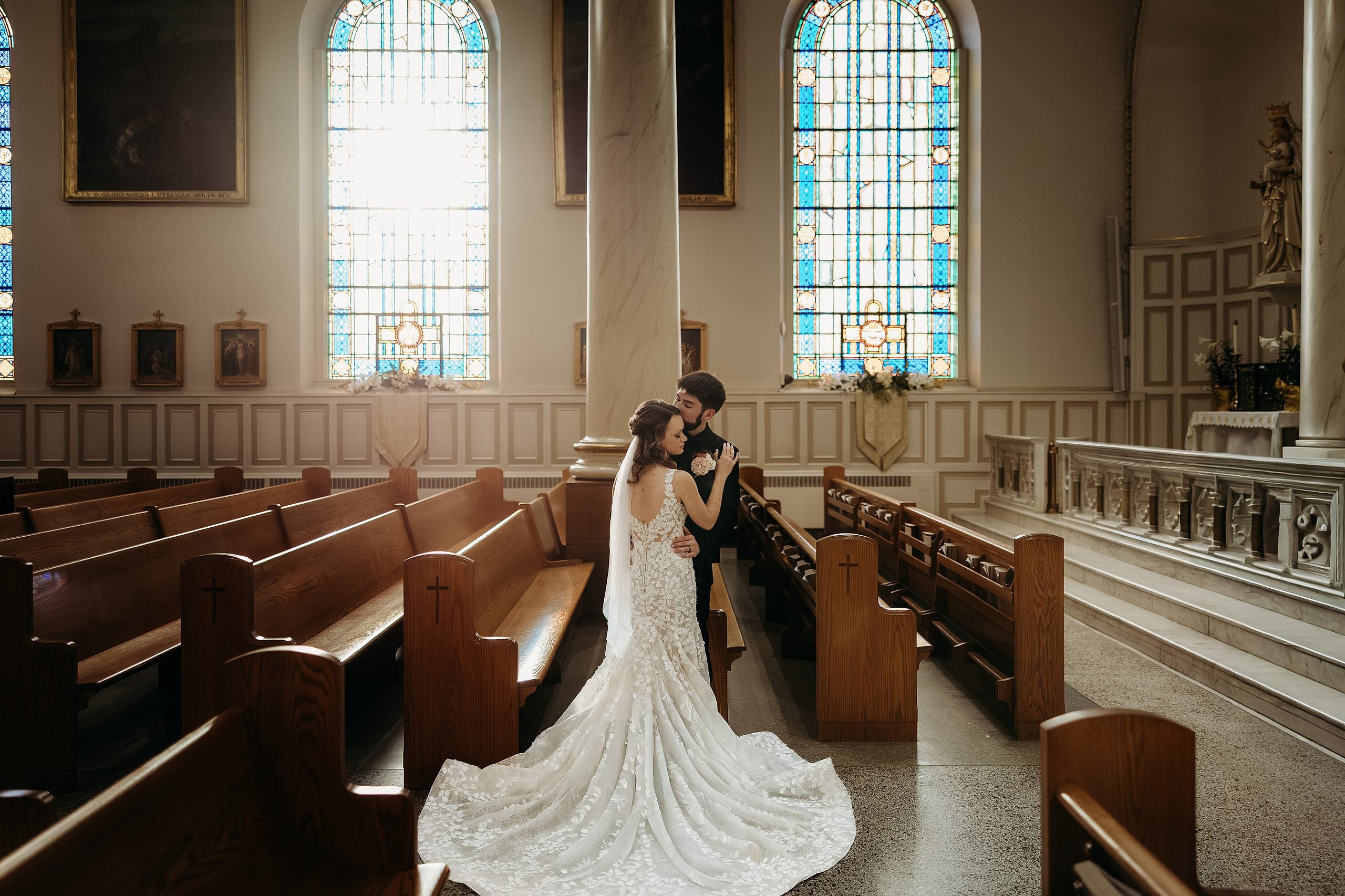 Bride and groom embrace inside Basilica of Saint Joseph beside glowing stained glass windows.
