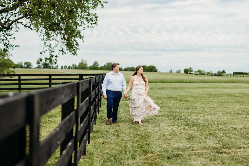 Engaged couple holding hands while walking beside a black fence in an open field.