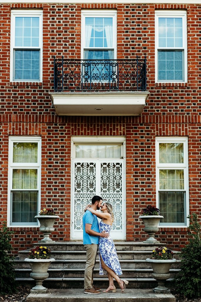 Engaged couple kissing on the steps of a brick estate surrounded by flowers.