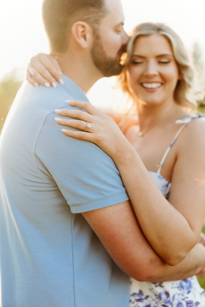 Close-up of an engagement ring as a smiling couple embraces in warm sunset light.