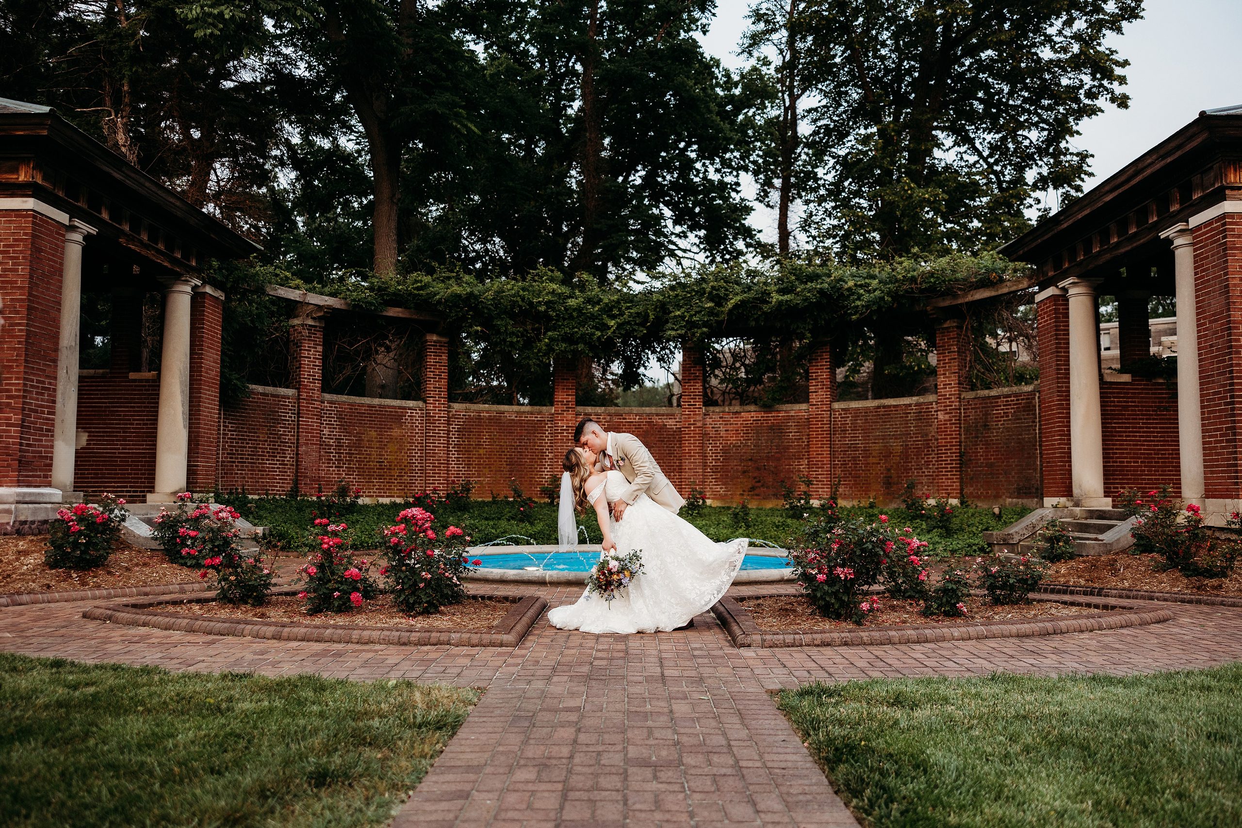 Bride and groom share a playful dip kiss in front of fountain surrounded by blooming roses and brick garden walls.