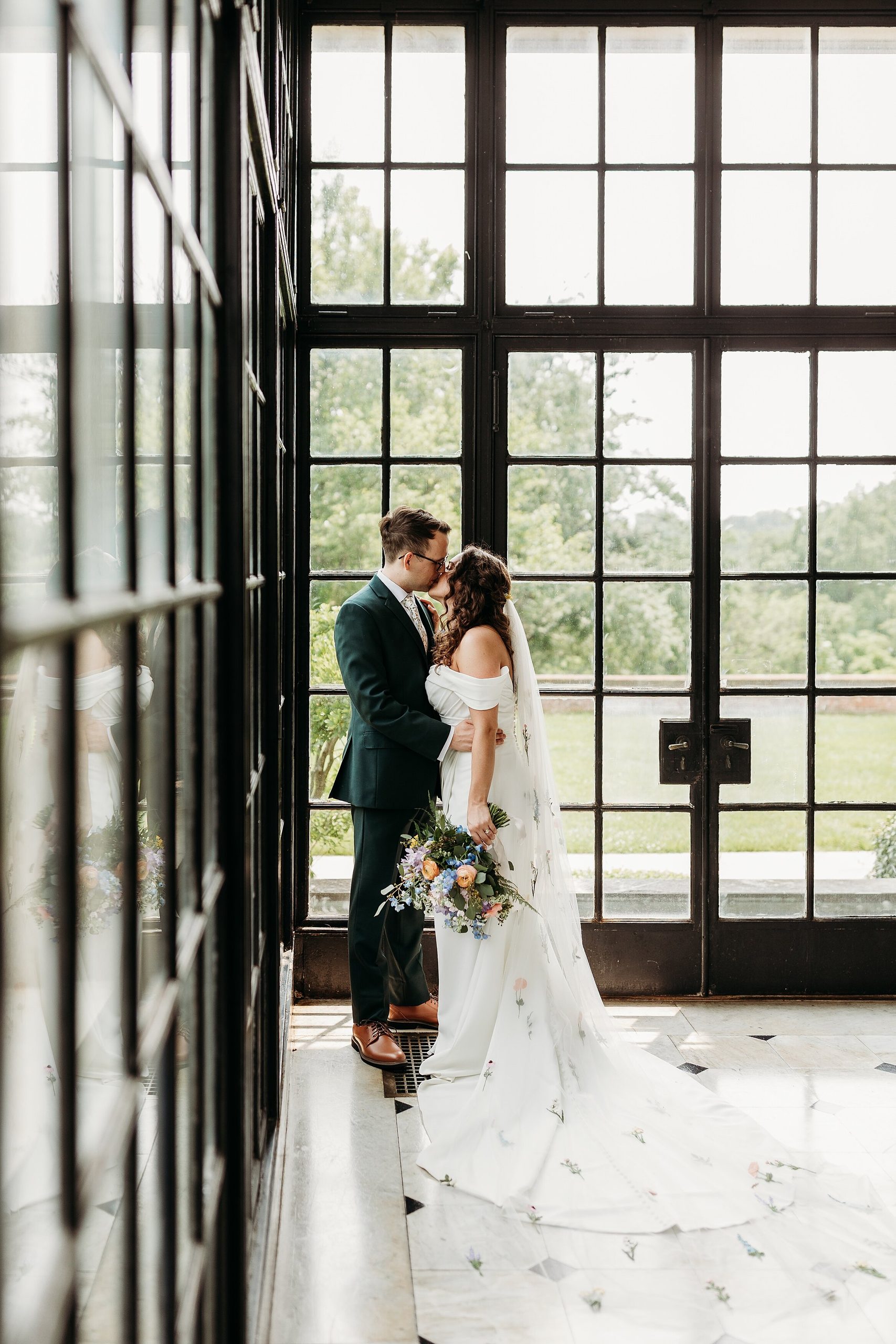 Couple kisses beside large windows during their Gardencourt wedding portrait.