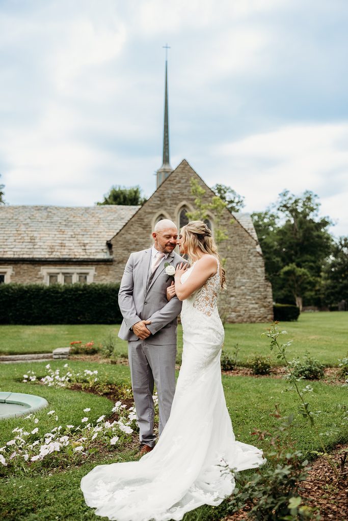 Bride and groom pose together in a garden with a stone chapel and steeple behind them.