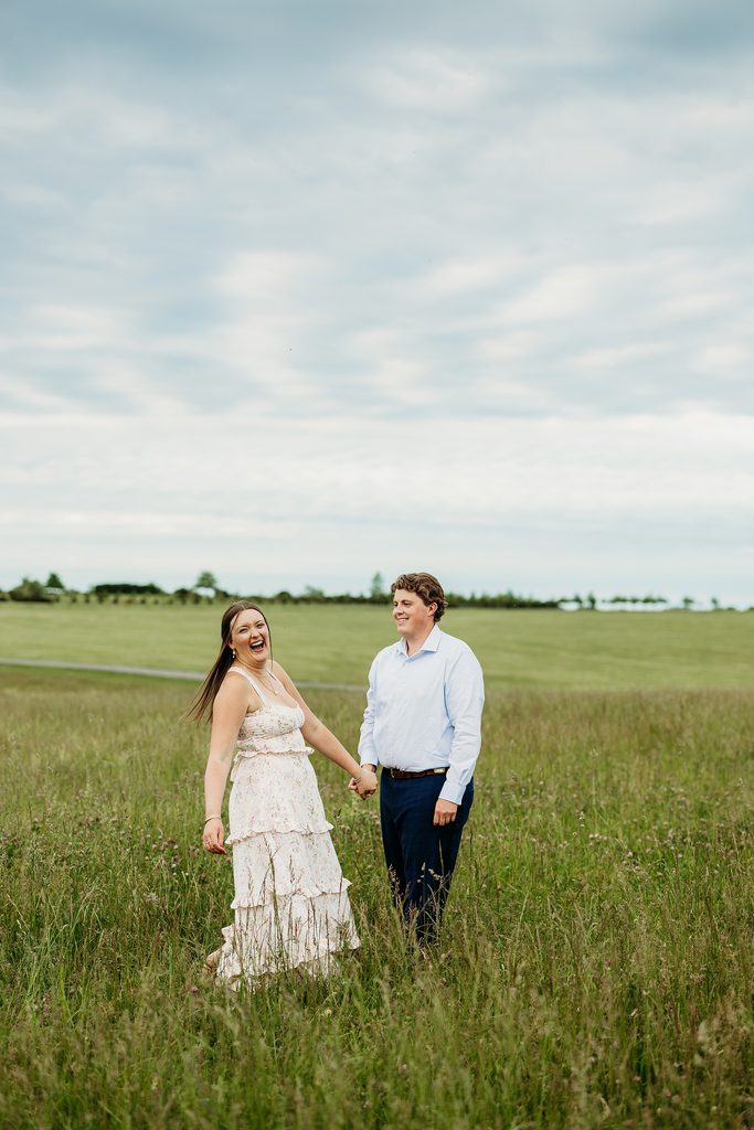 Engaged couple holding hands and laughing together in a grassy field.