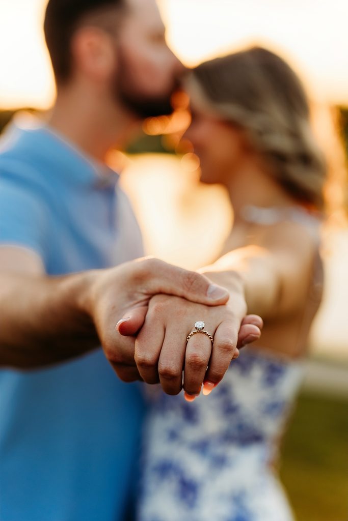 Engagement ring in focus as a couple holds hands and kisses at sunset.