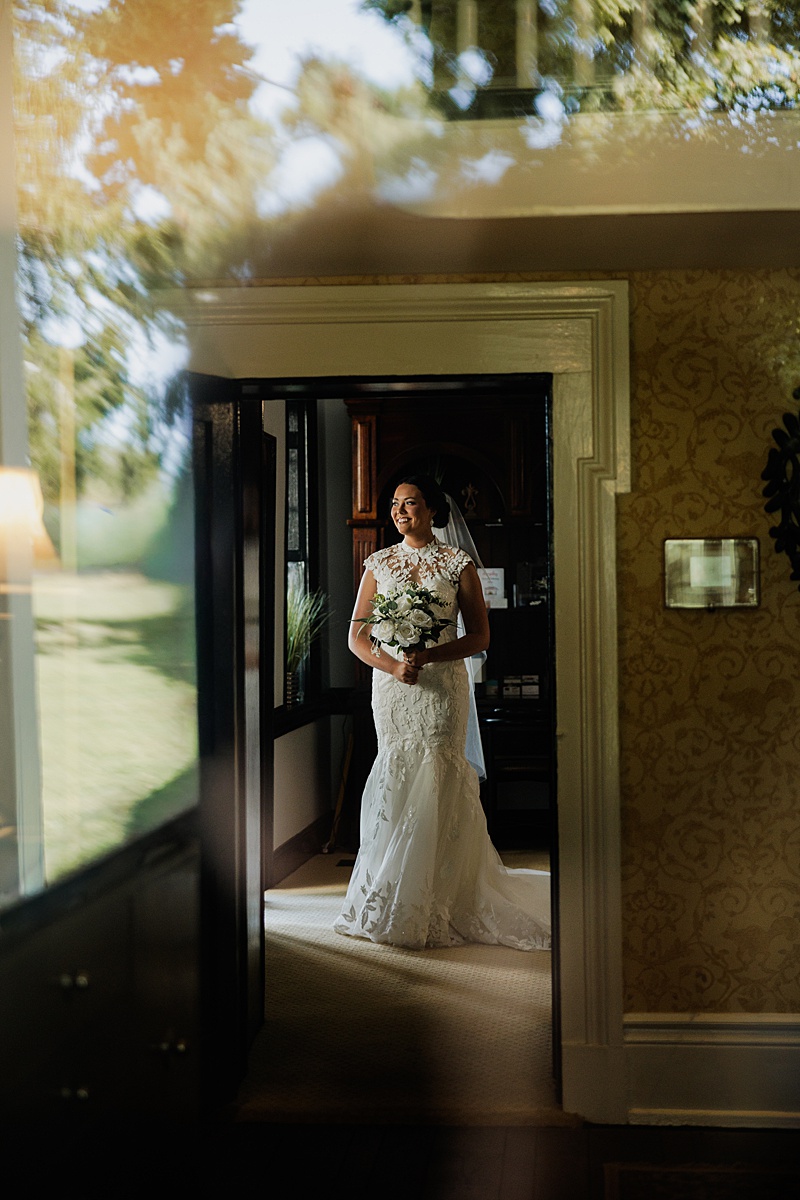 Bride holding a white bouquet framed by a doorway and window reflection.