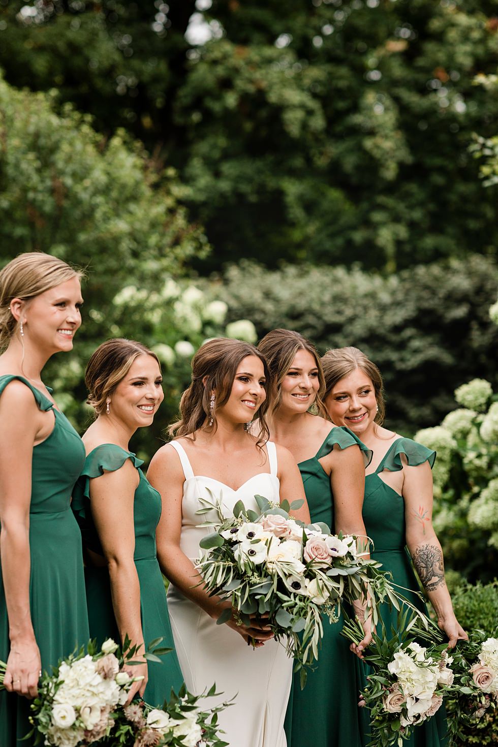 Bride poses with bridesmaids in green dresses surrounded by lush garden greenery.
