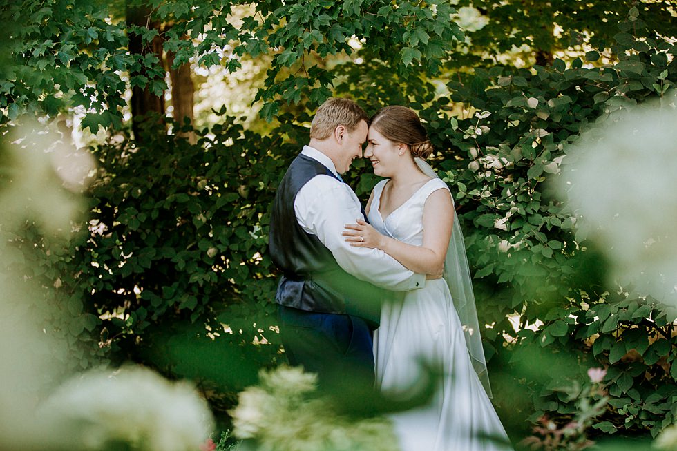 Bride and groom share a quiet moment surrounded by lush green foliage.